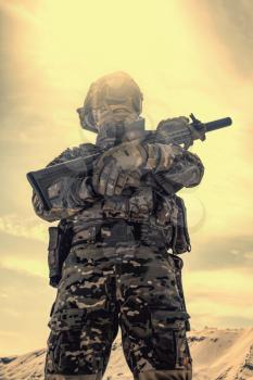 Airsoft player in U.S. army uniform, helmet, mask and glasses standing with service rifle replica in sandy area low angle, desaturated shoot. War games, military reenactment participant in desert