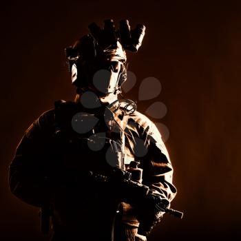 Army elite forces member, modern infantryman with hidden face, in tactical ammunition, equipped radio headset, night vision device mounted on helmet, standing with short barrel service rifle in hands
