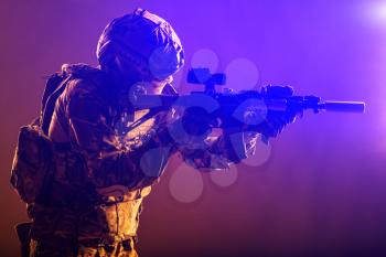 Modern soldier of army special forces, police anti-terrorist squad fighter in battle uniform, helmet with night vision device aiming short barrel assault rifle, low key studio shoot with red backlight
