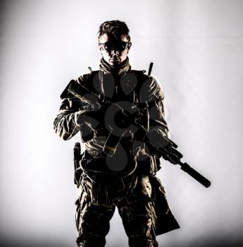 Military company mercenary, army special forces soldier, modern combatant in camouflage battle uniform, ballistic glasses, armed assault rifle with silencer low key studio portrait on white background