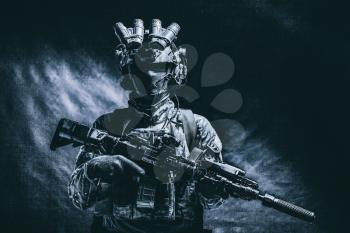 Half length, low key studio shoot of army soldier, marine infantryman in mask, camo uniform, equipped modern ammunition, armed service rifle standing in darkness with night vision device on helmet