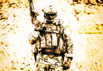 Army soldier, private military company fighter with hidden identity in battle uniform, helmet, body armor and radio headset, standing with service rifle in rocky area overexposed, desaturated shoot