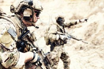 US special operations forces fighters armed with assault rifle, in opscore helmet, radio tactical ops headset, moving forward with caution in desert. Reconnaissance by fire in zone of war conflict