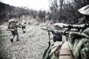 Modern warfare combatants group, commando fighters squad, team members attacking, rushing trough woodland. Sniper or marksman covering comrades in forest, over shoulder view