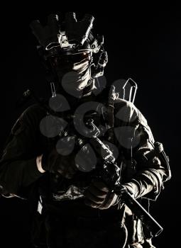 Army elite troops serviceman, counter-terrorist team member wearing mask and glasses, equipped night-vision device, radio headset mounted on combat helmet, armed submachine gun, standing in darkness