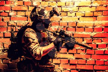 Anti-terrorist squad fighter, army elite forces soldier in combat uniform and tactical ammunition, armed mini submachine gun, wearing night-vision device, moving along brick wall during sqb operation