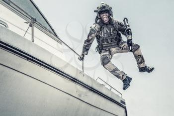Army commando, special forces soldier equipped with body armour and battle helmet with radio headset, screaming and yelling while jumping from speed boat deck, landing on shore with assault rifle