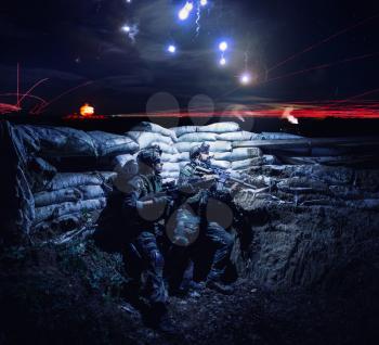 Army soldiers armed assault rifles, hiding in trench during night action. Navy SEALS team fighters, commando shooters with weapon, watching on falling flares in sky, waiting for signal to attack