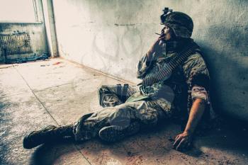 Soldier in combat uniform and ammo belt over chest, sitting on floor and smoking after being shoot in shoulder and put bandage on wounded hand, dropped weapon lying nearby with blood stains around