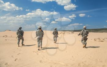 Team of United states airborne infantry men with weapons moving patrolling desert. Sand and blue sky on background of squad, sunlight, back view