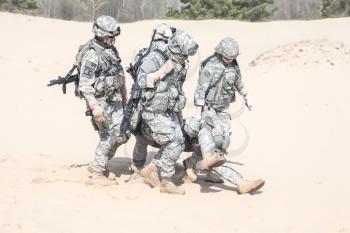 Team of airborne infantry paratroopers saving life of injured brother in arms dragging carrying him on desert sand. No man left behind