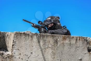 Swat police operator with sniper rifle in black uniforms aiming criminals terrorists waiting in stakeout behind concrete block. Sunny day, low angle