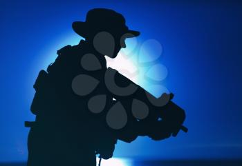 Silhouette of army special forces commando, soldier in boonie hat aiming and shooting bullpup submachine gun on background of setting sun. Private military company mercenary engaging enemy at sunset