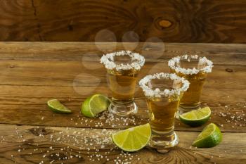 Three shots of gold tequila with lime and salt on the wooden background. Tequila shot. Gold Mexican tequila. Tequila