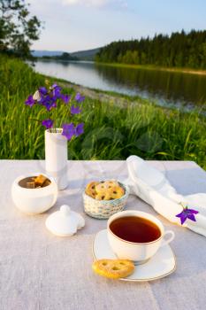 Evening tea sunset river view. Cup of tea and cookies in small wicker basket with blue bell flowers in vase. Summer tea time concept. 