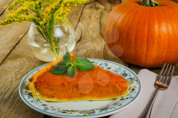 Thanksgiving pumpkin pie slice on white plate, linen napkin, vase, yellow flowers on a wooden background
