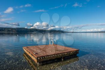 A wooden swim float sits on Hood Canal in Washington State. Blue cloudy skies are reflected in calm waters.