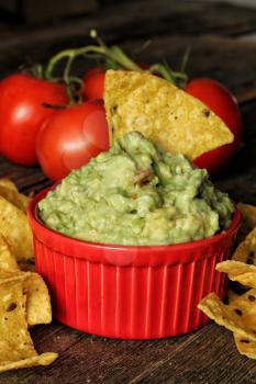 Guacamole with nachos in a red bowl with fresh tomato in background