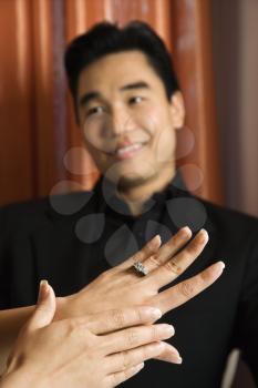 Prime adult Asian female admiring engagement ring with male in background.