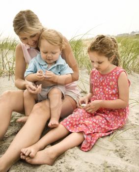 Caucasian mid-adult woman sitting with male toddler on lap and beside Caucasian female child on beach  looking at shells.