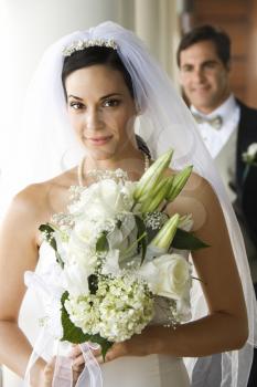 Caucasian mid-adult bride holding bouquet with groom in background.