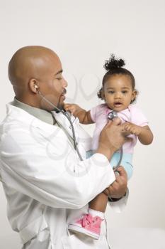 African-American male pediatrician holding and examinating baby girl with stethoscope.