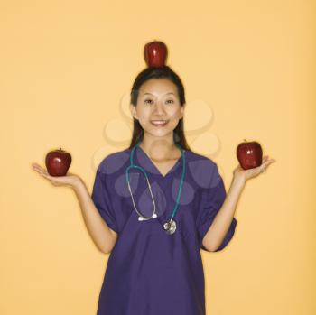 Royalty Free Photo of a Female Doctor Holding Two Red Apples in Hands and Balancing One on Her Head