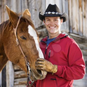 Royalty Free Photo of a Young Caucasian man in cowboy hat petting his horse