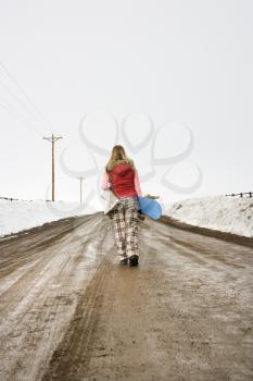 Royalty Free Photo of a Woman Wearing Winter Clothes Walking Alone on a Muddy Dirt Road Holding a Snowboard and Boots