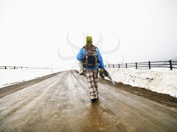 Royalty Free Photo of a Woman Wearing Winter Clothes Walking on a Muddy Dirt Road Holding a Snowboard and Boots