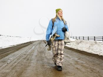 Royalty Free Photo of a Woman in Winter Clothes Walking Down Muddy Dirt Road Holding Snowboard and Boots Smiling