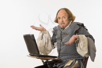 Royalty Free Photo of William Shakespeare in Period Clothing Sitting in a School Desk Shrugging at a Laptop