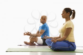 Side view of mid adult multiethnic man and woman sitting in lotus position on exercise mats with eyes closed and legs crossed.