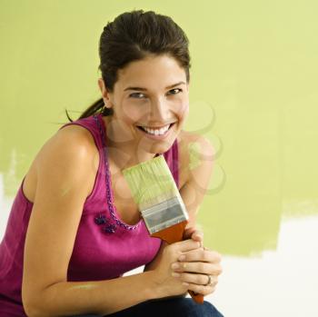Royalty Free Photo of a Smiling Woman Kneeling in Front of a Partially Painted Wall Holding a Paintbrush