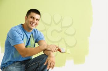 Royalty Free Photo of a Smiling Man Kneeling in Front of a Partially Painted Wall Holding a Paintbrush