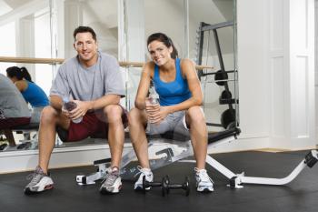 Man and woman sitting on exercise machine talking holding water bottles.