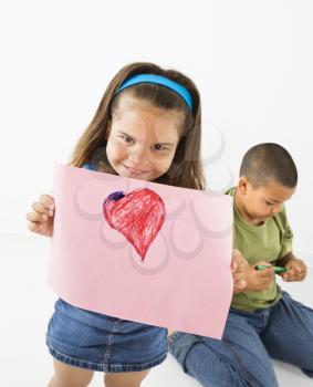 Royalty Free Photo of a Girl Showing Off a Drawing of a Heart While a Boy Sits Behind Ber