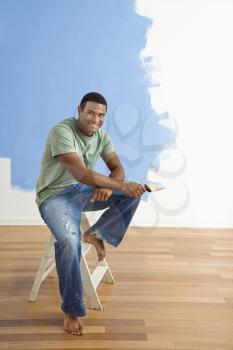 Royalty Free Photo of a Young Man Sitting Next to a Half-Painted Wall Holding a Paintbrush