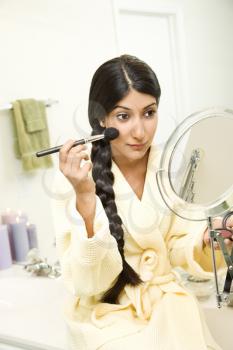A young woman wearing a bathrobe is sitting on the bathtub  in front of a mirror and applying makeup. Her long dark hair is braided and hanging over her shoulder. Vertical shot.