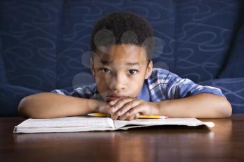 Young African American boy sits on the floor at a coffee table. He is looking towards the camera with his homework on the table. Horizontal shot.