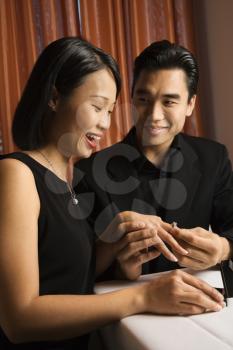 Attractive young Asian man places an engagement ring on an attractive young Asian woman. Vertical shot.