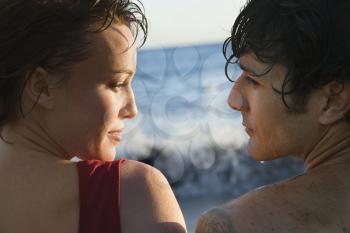 Cropped close-up of young man and woman, wet from swimming, looking at each other, with the ocean in the background. Horizontal shot.