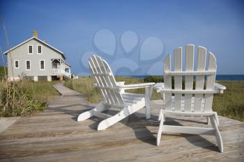 Two Adirondack style chairs sitting on a wooden deck, facing the shore. There is a large home in the background. Horizontal shot.