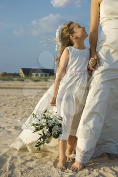 Flower girl looks up towards a bride on a sandy beach. Vertical shot.