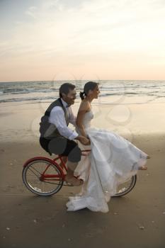Bride rides the handle bars of a bicycle being driven by her groom on the beach. Vertical shot.