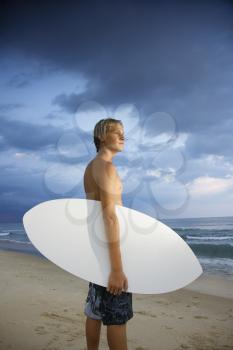 Young male surfer standing on beach with surfboard looking out at ocean.