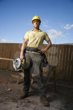 Worker in a hardhat and safety glasses stands at a construction site with a hand on his hip. He holds a circular saw with the other hand. Vertical shot.