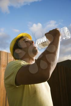 Side view of a male Caucasian construction worker as he drinks from a plastic water bottle. A fence and the blue sky can be seen in the background. Vertical shot.
