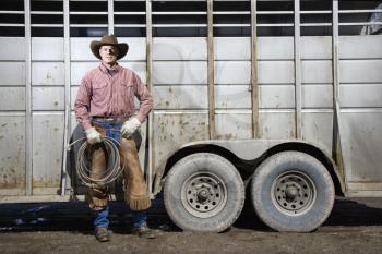 Man wearing a cowboy hat, leaning on the side of a livestock trailer. He is holding a lariat. Horizontal shot.