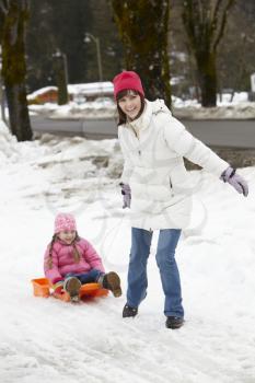 Mother Pulling Daughter On Sledge Along Snowy Street In Ski Resort